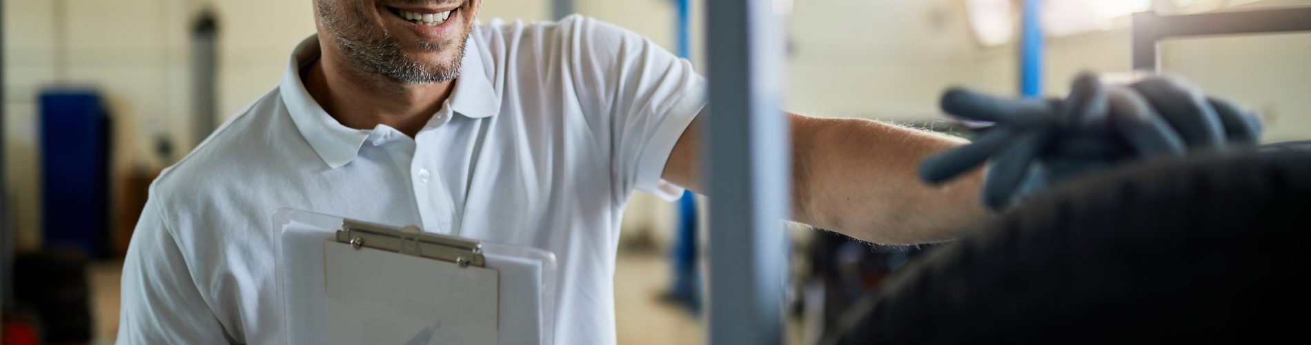 Uma foto horizontal mostra um gestor de frota sorrindo com um boné azul e camisa polo branca, segurando uma prancheta enquanto aponta para o pneu de um carro. Ele tem barba e está olhando para a direita. No fundo, há uma oficina mecânica de borracha e metal. Uma janela à direita brilha com a luz do sol, o pneu do carro está desfocado em primeiro plano, mas a mão direita do mecânico está sobre ele. Sua mão esquerda segura a prancheta branca com a caneta azul.